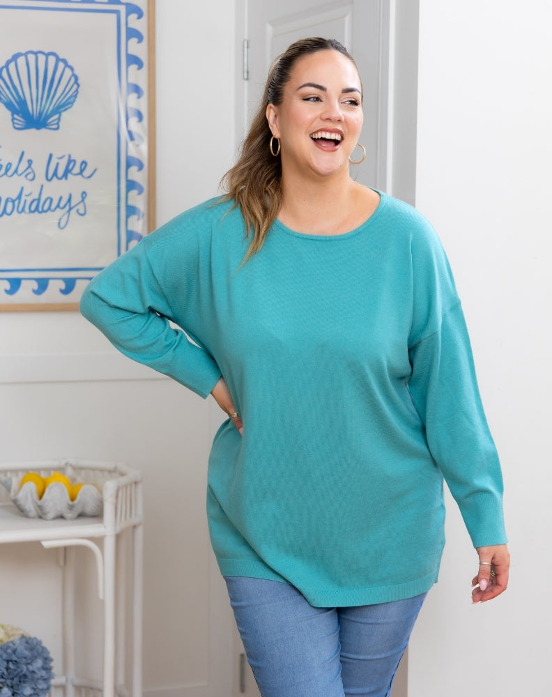 Woman wearing a teal sweater and blue jeans standing in a room with a decorative wall sign.