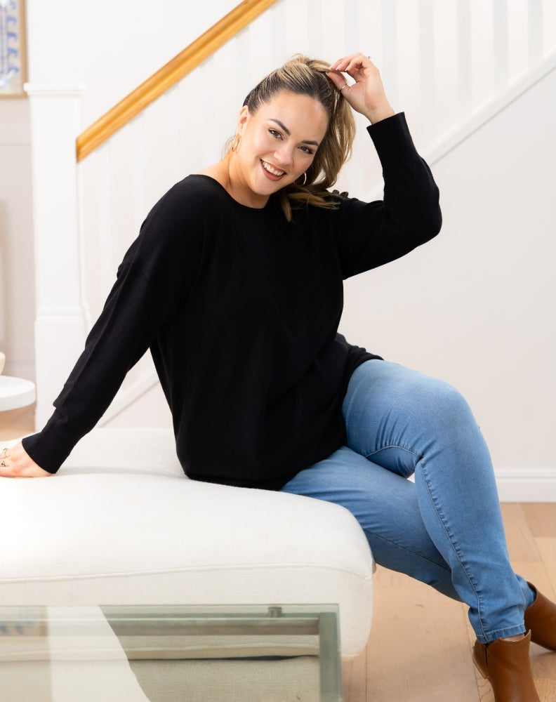 Woman sitting on a white stool wearing a black tunic knit and blue jeans in a bright room.