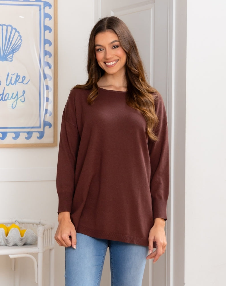 Woman wearing a brown long-sleeve top and blue jeans standing in a room with a decorative wall piece.