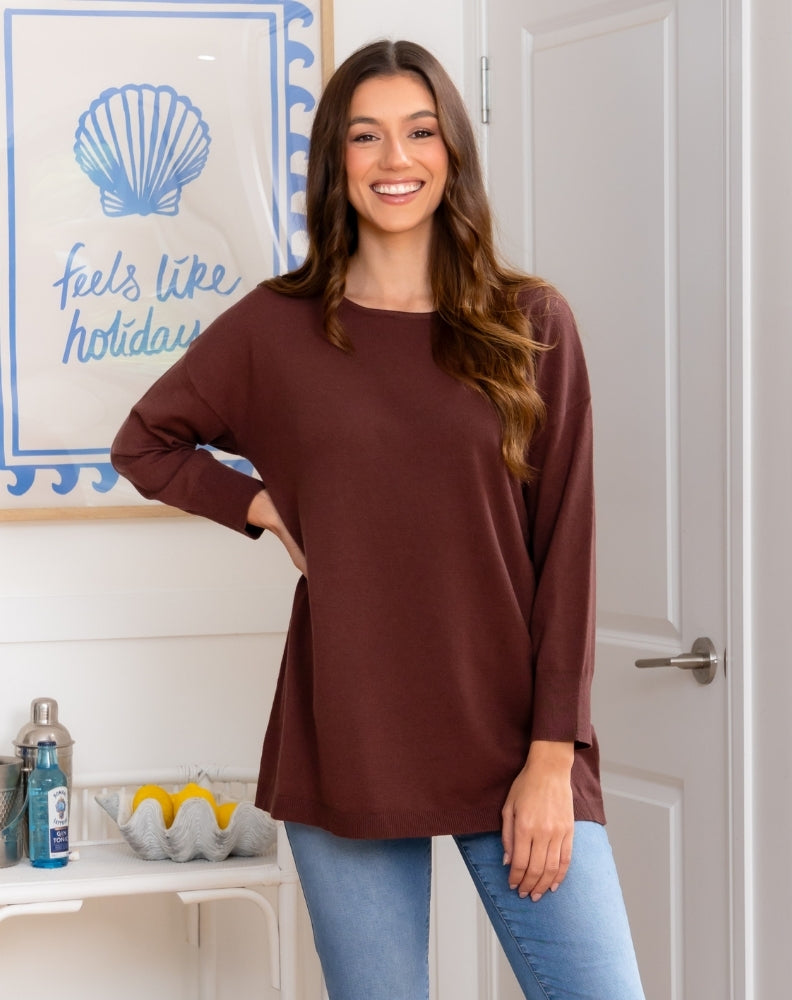Woman wearing a brown long-sleeve top and blue jeans standing in a room with a decorative wall sign.