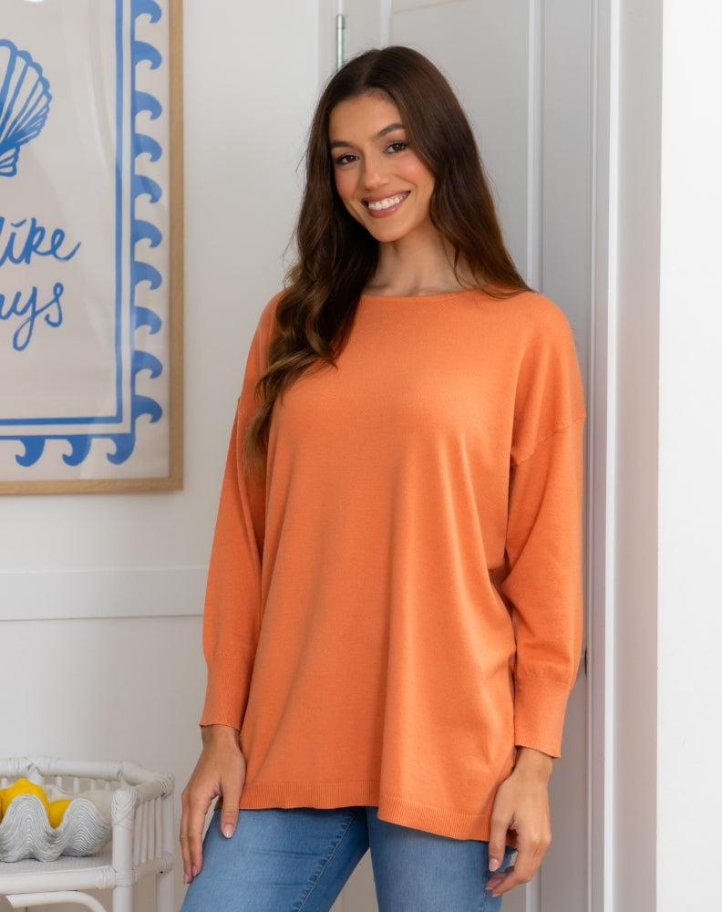 Woman wearing an orange long-sleeve top and blue jeans standing in a room with a decorative wall hanging.