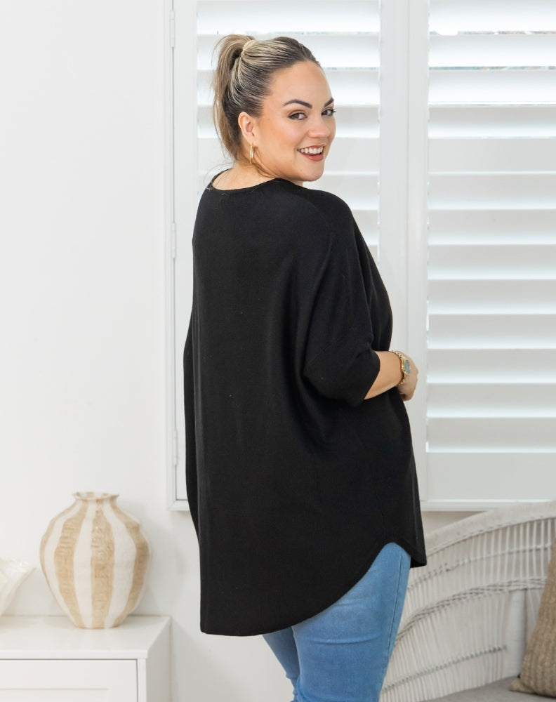 A woman wearing a black long-sleeve tunic top and blue jeans, standing in a bright room with white shutters and a decorative vase.
