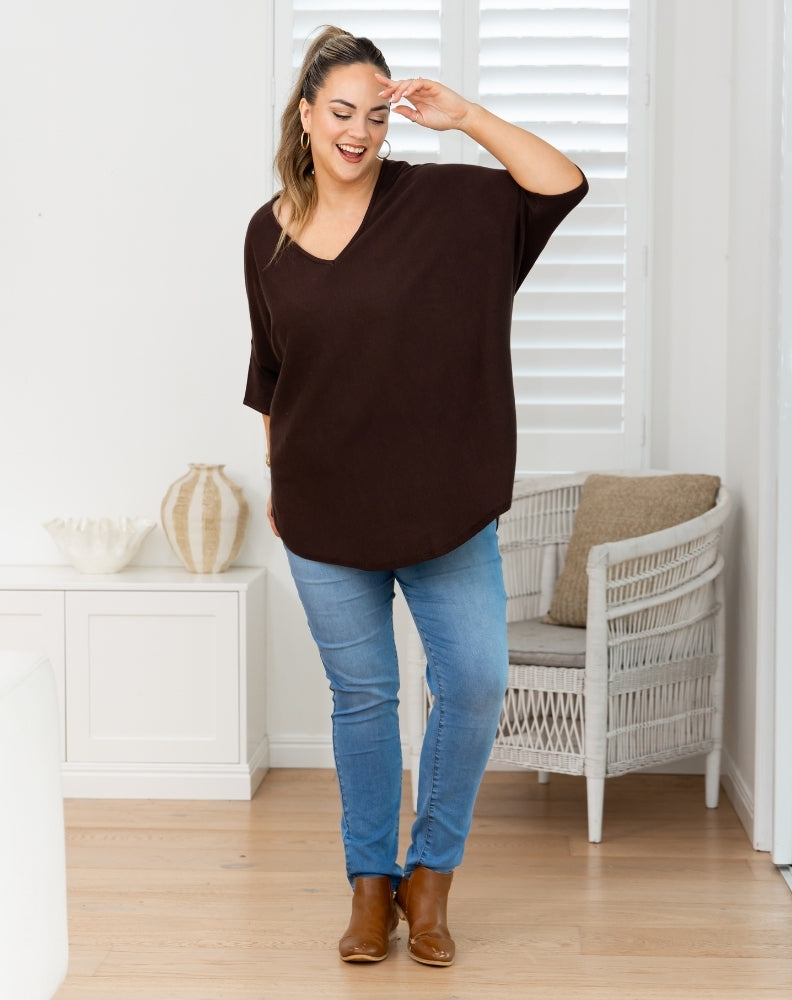 A woman modeling a dark brown oversized short-sleeve V-neck top with blue jeans and brown ankle boots, standing in a bright room with a white chair and window blinds.