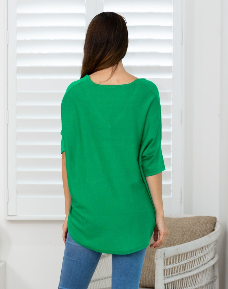 A woman viewed from the back wearing a bright green, loose-fitting short-sleeve top and blue jeans, standing in front of white window shutters.