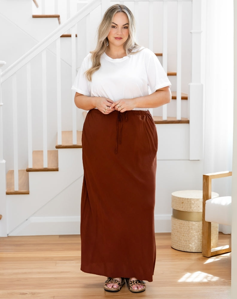 Woman wearing a white top and brown skirt standing in a home setting with stairs in the background.