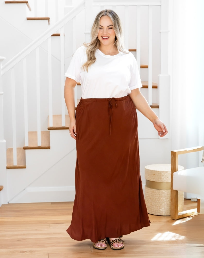 Woman wearing a white top and brown maxi skirt standing in a home setting.