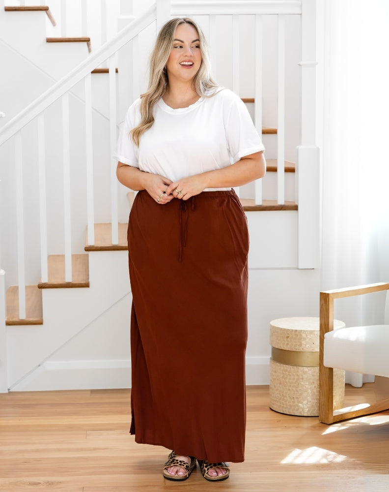 Woman wearing a white top and brown skirt standing in a home setting with stairs in the background.