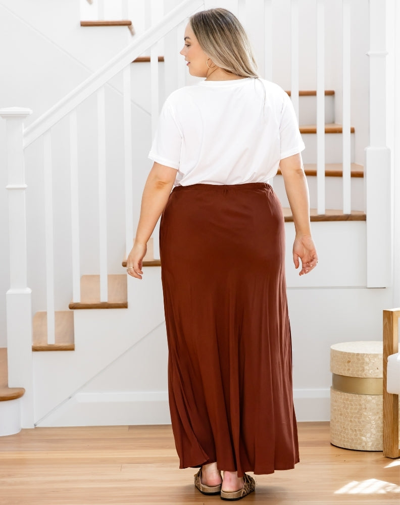 Person wearing a white top and brown skirt standing in a home setting with stairs in the background.