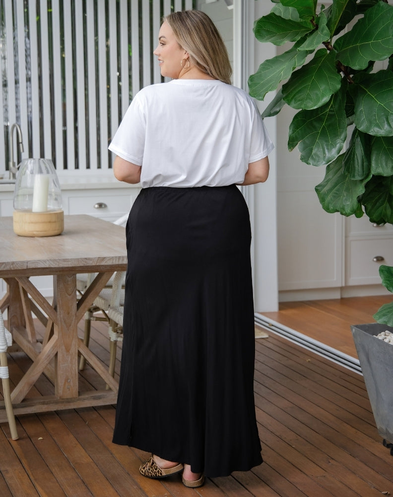 Woman wearing a white top and black skirt standing on a wooden deck with plants and furniture in the background.