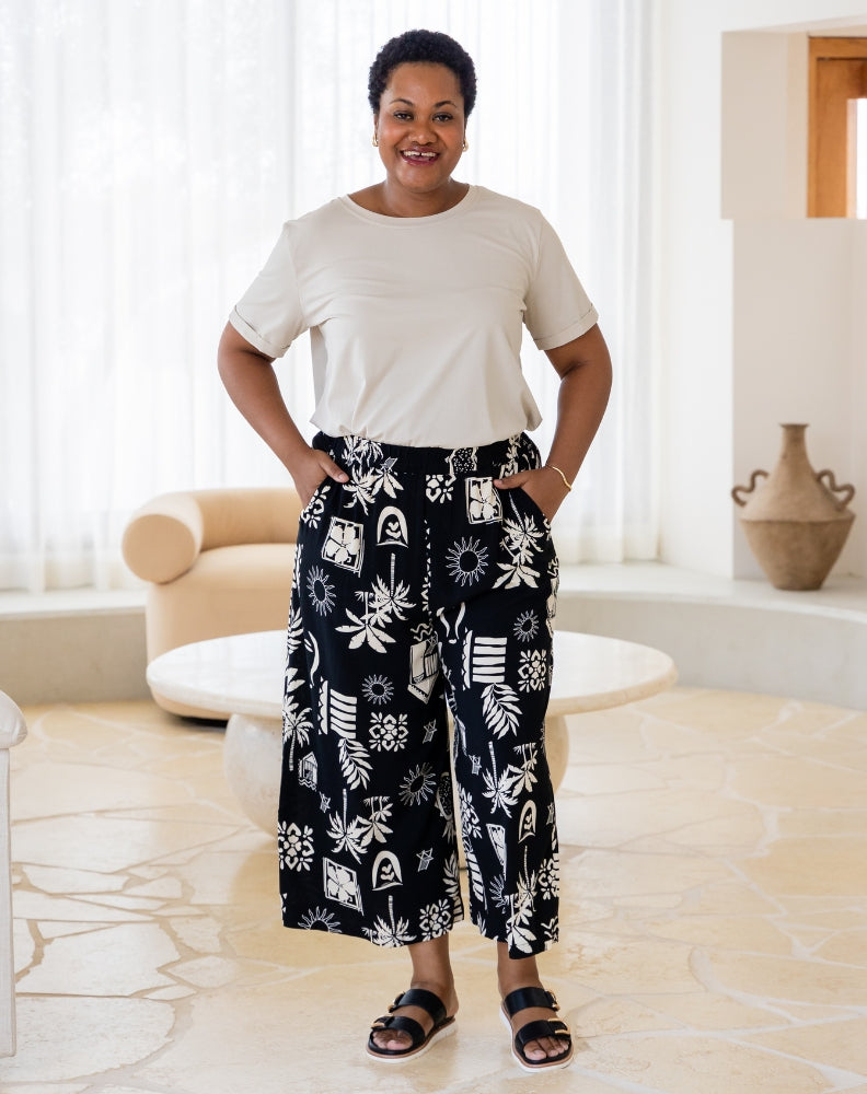 Woman wearing a white top and black patterned pants in a room with a couch and vase.