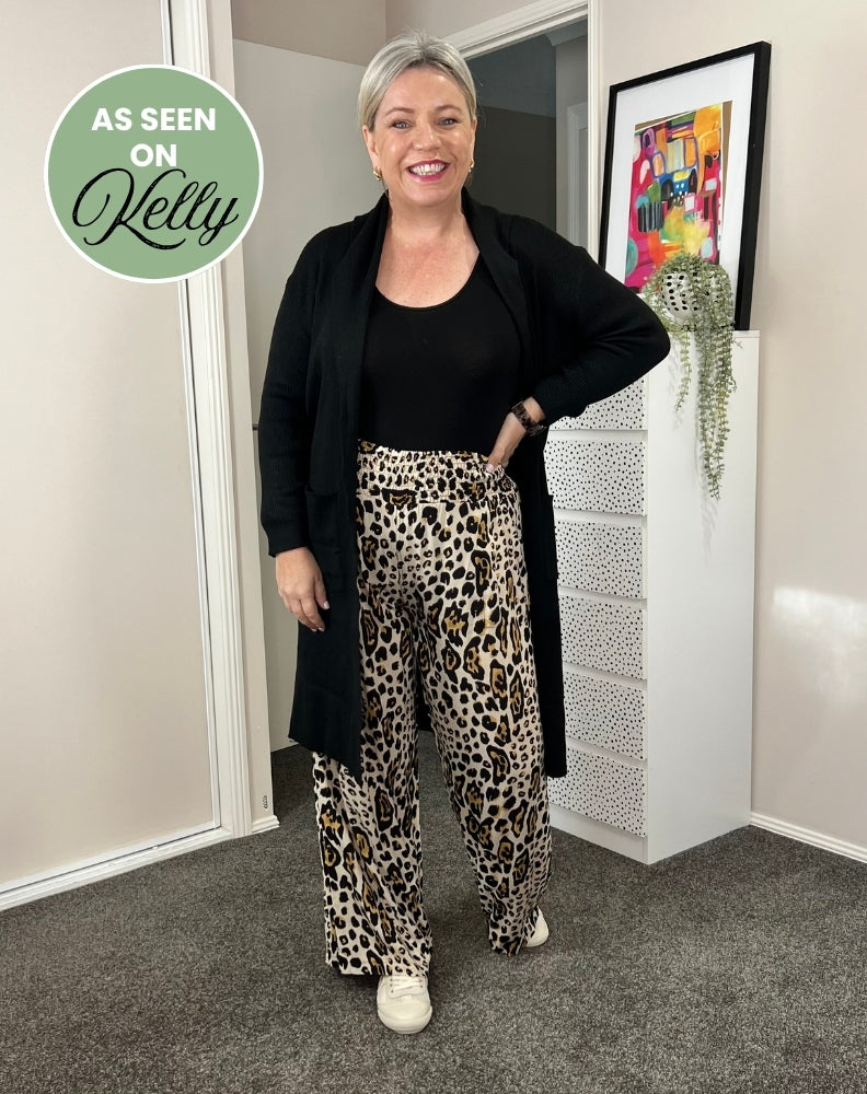 Woman wearing a black cardigan, black top, and leopard print pants in a room with a colorful painting on the wall.
