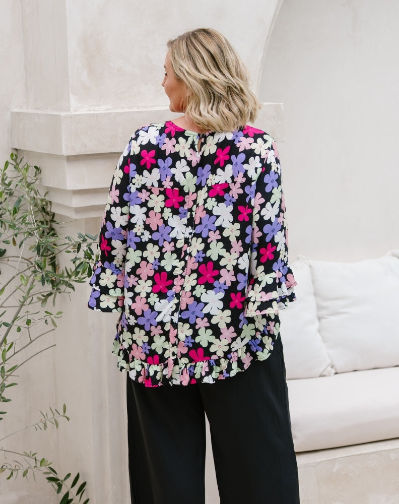 A woman shown from the back wearing a black blouse with a colorful floral pattern in pink, purple, and white, paired with black wide-leg pants, standing in a bright, modern interior setting.