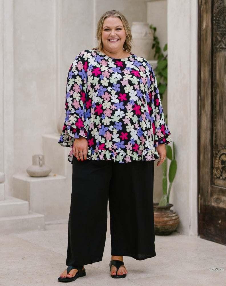 A woman modeling a long-sleeve floral blouse with black wide-leg pants and black sandals, standing in a light-colored stone doorway with plants in the background.