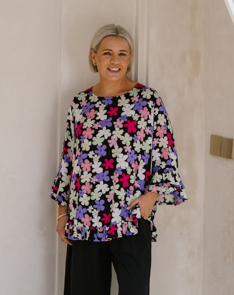 A woman with short blonde hair wearing a black blouse with a colorful floral pattern in pink, purple, and white, paired with black trousers, standing against a beige wall.