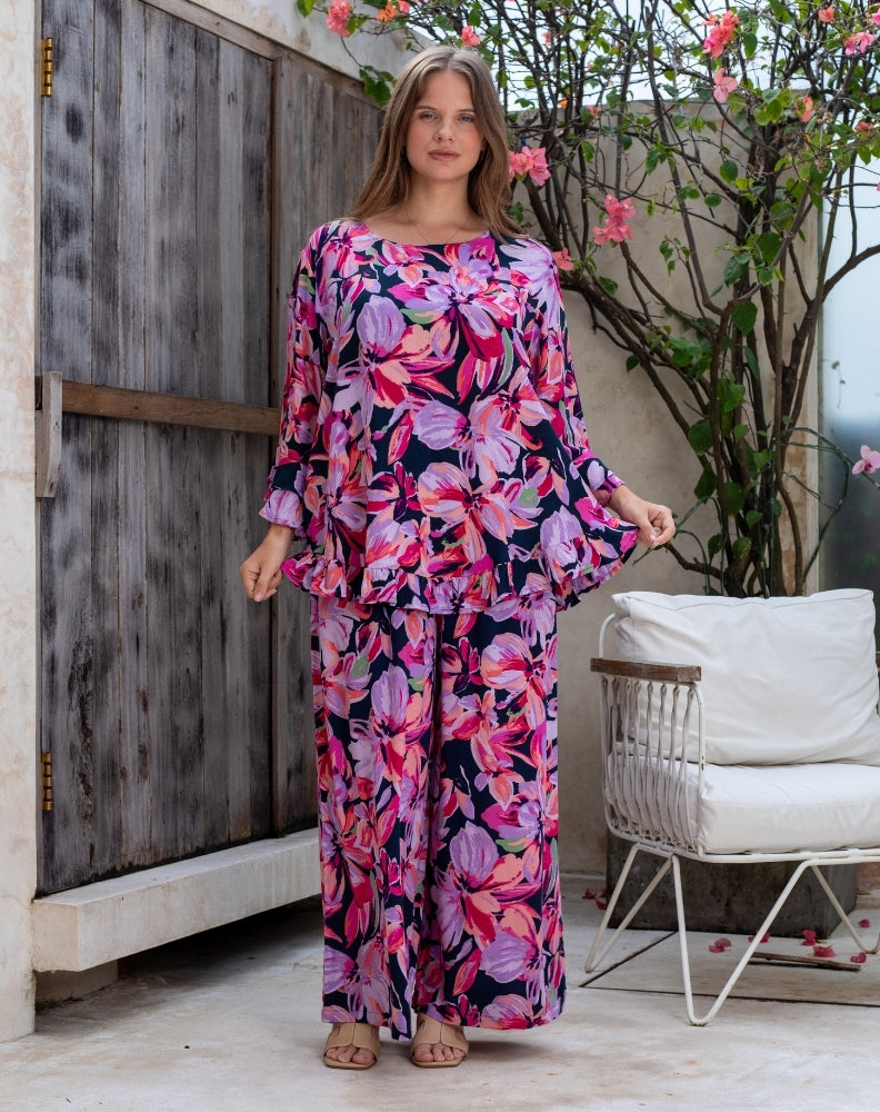Woman wearing a colorful floral dress standing in an outdoor setting with a wooden door and white chair.