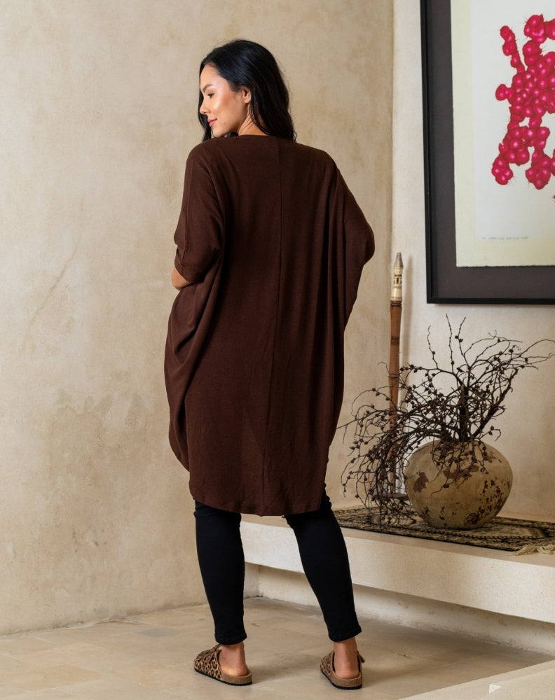 Woman wearing a brown dress standing in a room with a decorative vase and artwork on the wall.