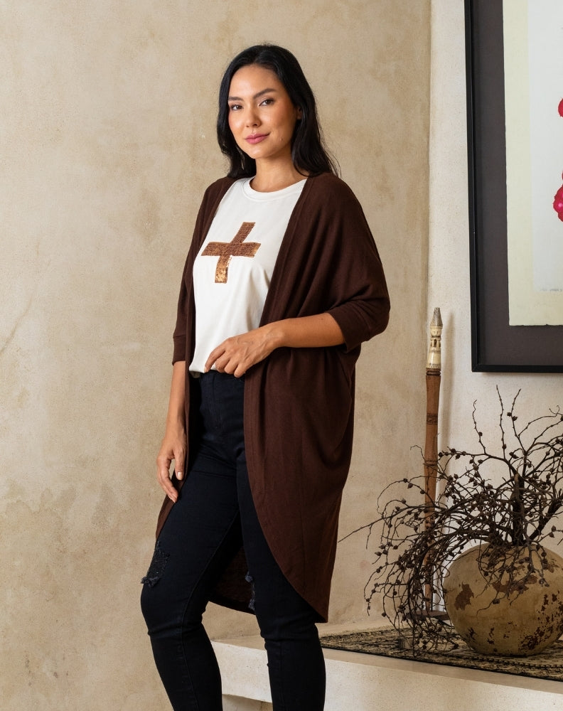 Woman wearing a brown cardigan over a white shirt with a logo, standing indoors.