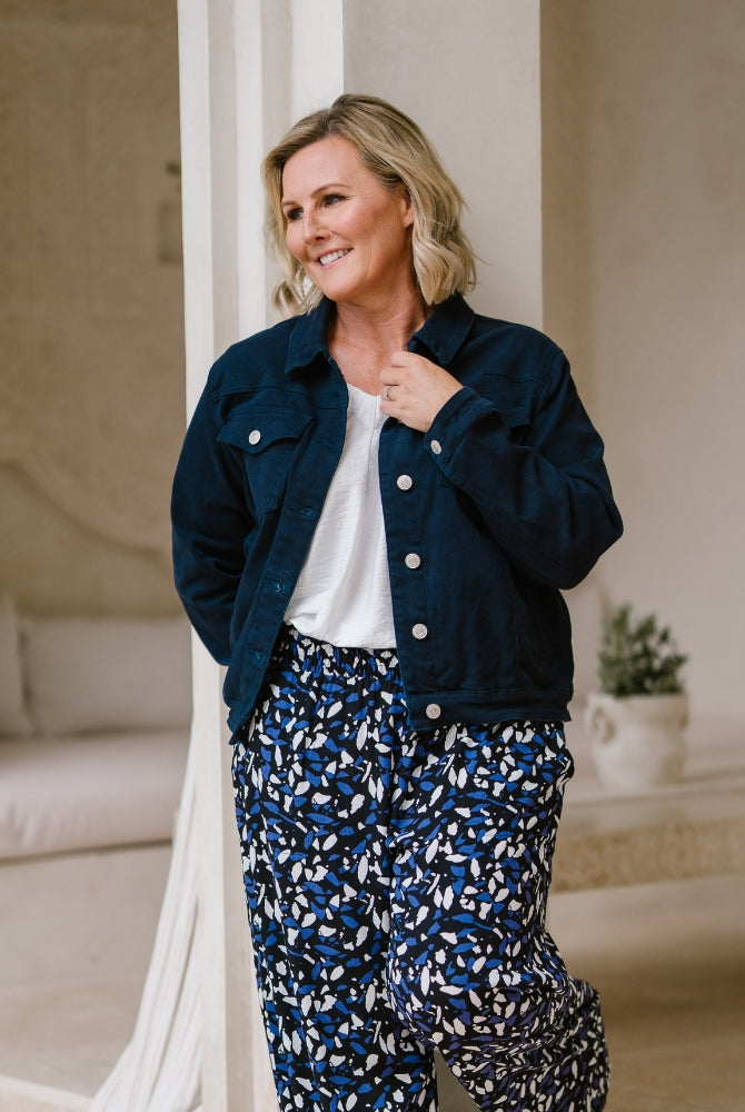 A woman wearing a dark navy blue denim jacket over a white top and blue-and-white patterned pants, standing in a softly lit indoor setting with neutral walls and furniture.