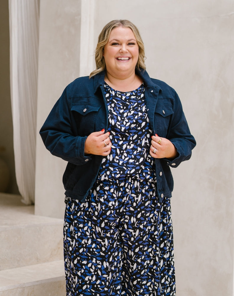A woman smiling and wearing a navy blue denim jacket over a matching navy, white, and blue patterned jumpsuit, standing in front of a neutral beige wall.