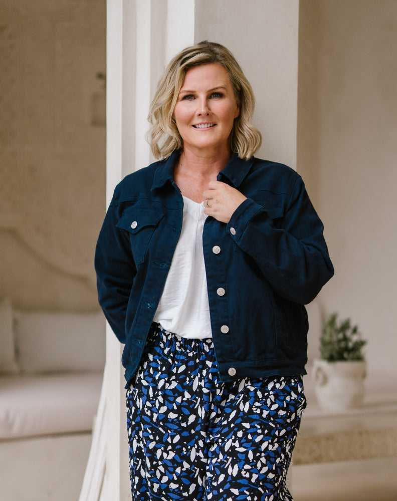 A woman modeling a dark navy blue button-up jacket over a white t-shirt and blue-and-white patterned pants, standing in a softly lit indoor setting with neutral-toned walls and a small potted plant in the background.
