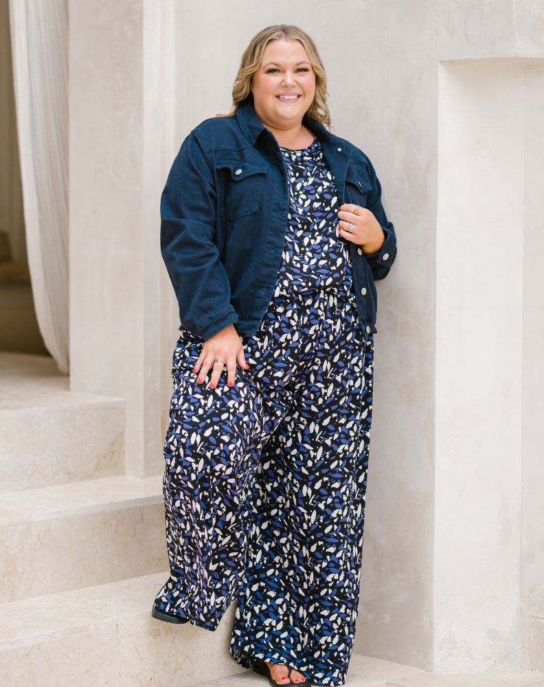 A woman stands on stone steps wearing a navy blue denim jacket over a matching navy, white, and blue abstract print jumpsuit.