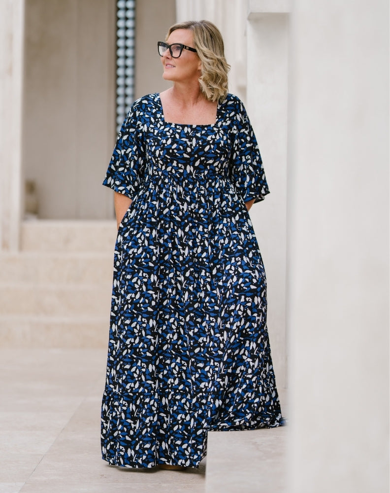 A woman wearing a navy blue maxi dress with a white and light blue abstract floral pattern, standing in a bright, modern outdoor corridor.