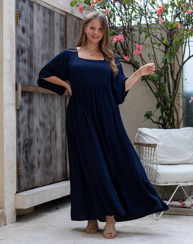 Woman wearing a navy blue dress standing outdoors near a wooden wall and white chair.