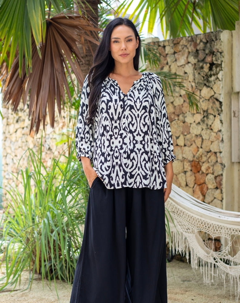 Woman wearing a black and white patterned top and black pants standing in a tropical setting with plants and a stone wall.