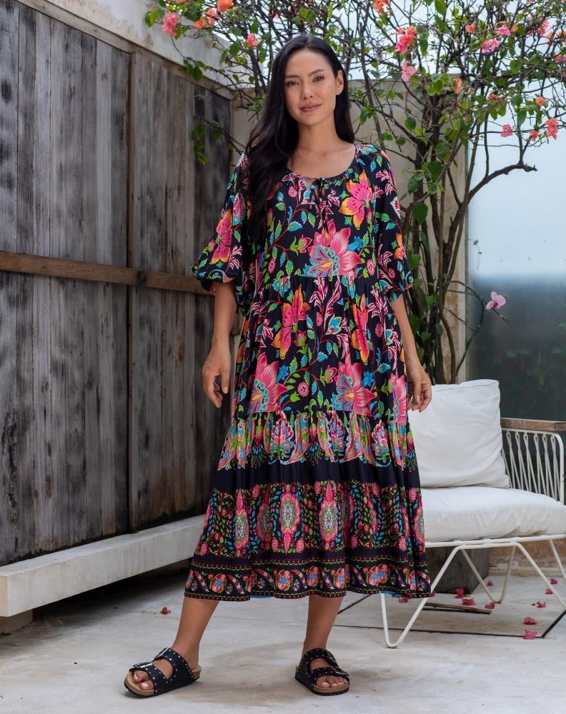 Woman wearing a colorful floral dress standing outdoors with a wooden wall and white chair in the background.