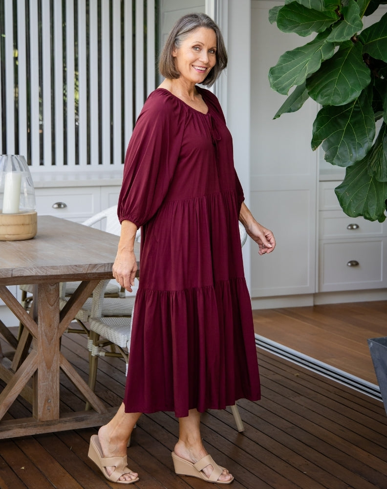 Woman in a burgundy dress standing on a wooden deck with a plant and table in the background.