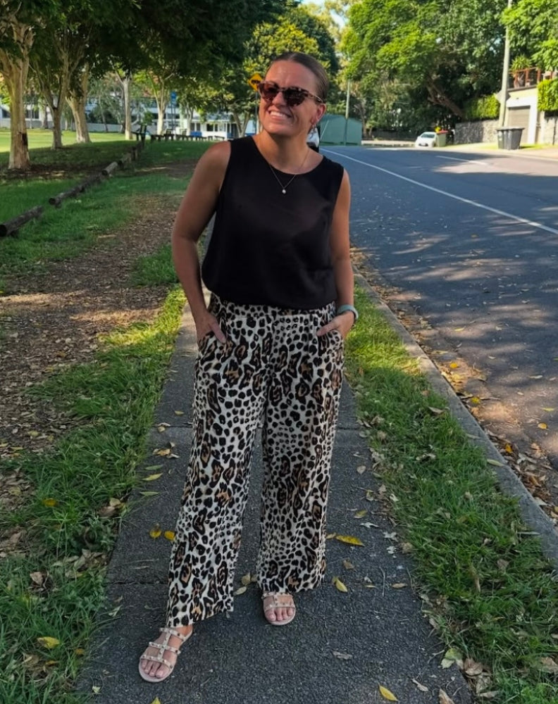 Woman wearing a black sleeveless top and leopard print pants standing on a sidewalk with trees and a road in the background.