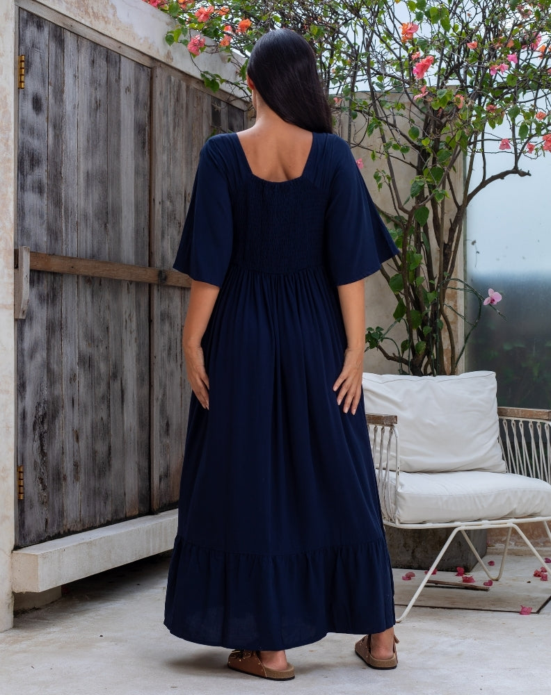 Woman in a navy blue dress standing outdoors near a wooden fence and white chair.
