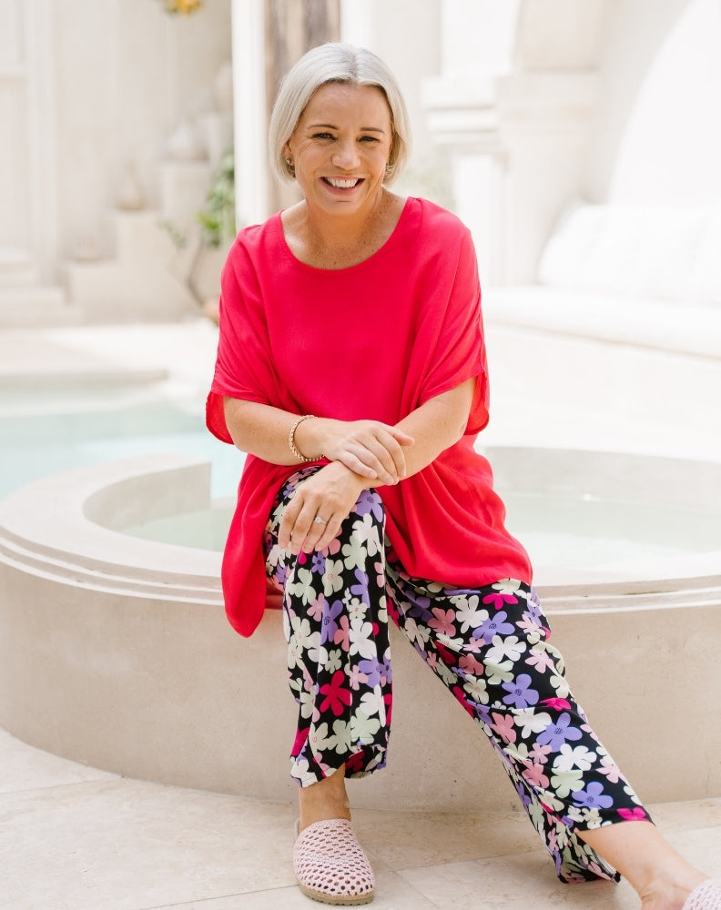 A smiling woman with short gray hair sits on the edge of a light-colored stone fountain. She is wearing a bright coral-red short-sleeve tunic top and black wide-leg pants with a colorful floral print in pink, purple, white, and green.