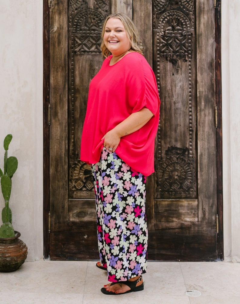 A woman modeling a bright pink short-sleeve top and black wide-leg pants with a colorful floral pattern, standing in front of a large dark wooden door.