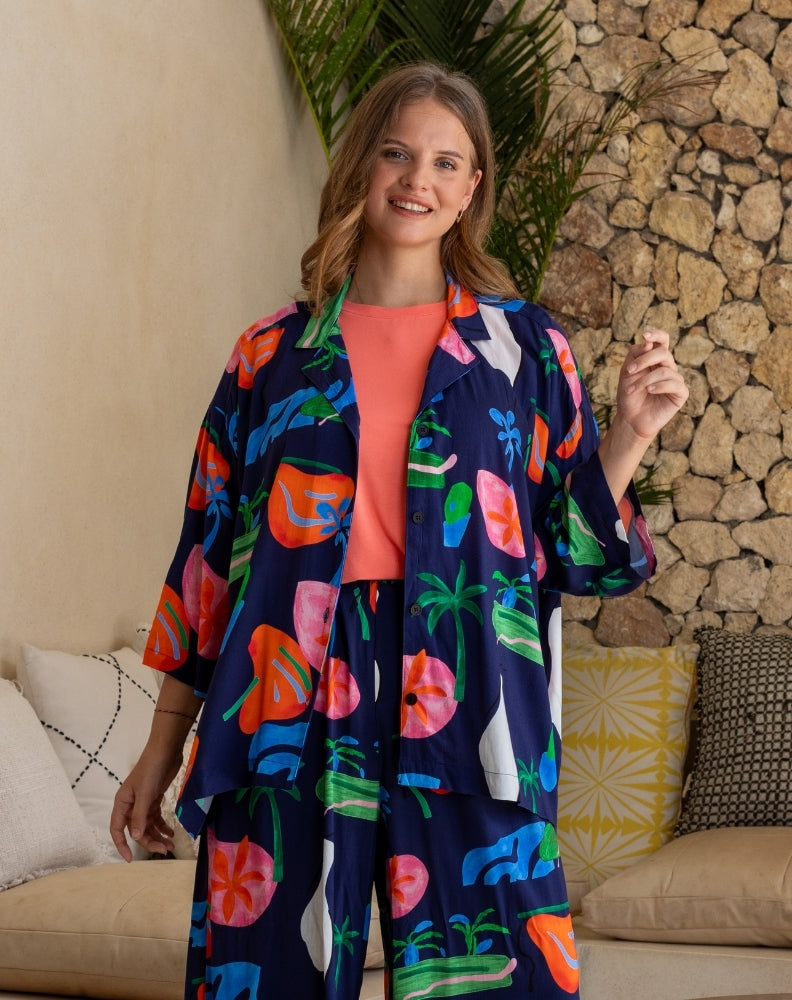 Woman wearing a colorful kimono-style outfit in a casual setting with a stone wall and plants.