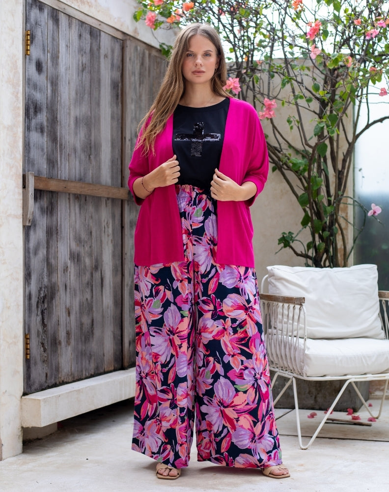 Woman wearing a bright pink cardigan over a black top and floral pants in an outdoor setting.
