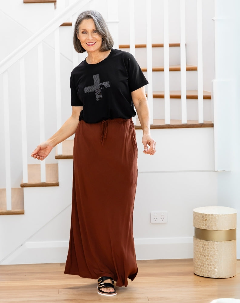 Woman wearing a black t-shirt with a graphic design and a long brown skirt, standing in a home setting.