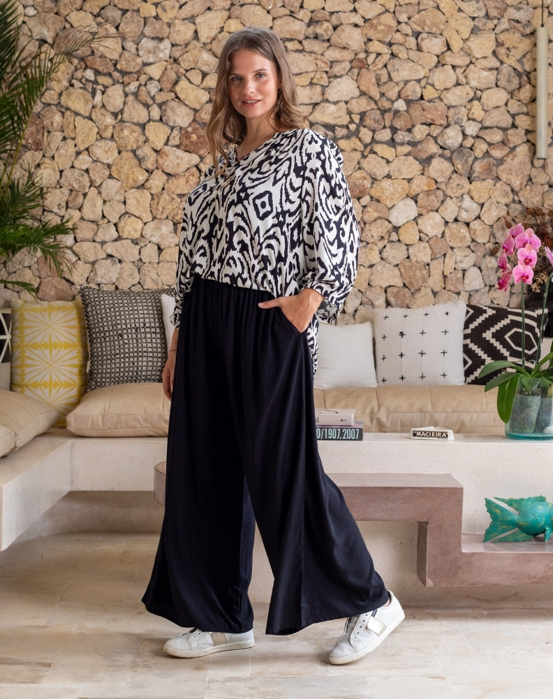 Woman in a patterned top and black pants standing in a living room with a stone wall background.