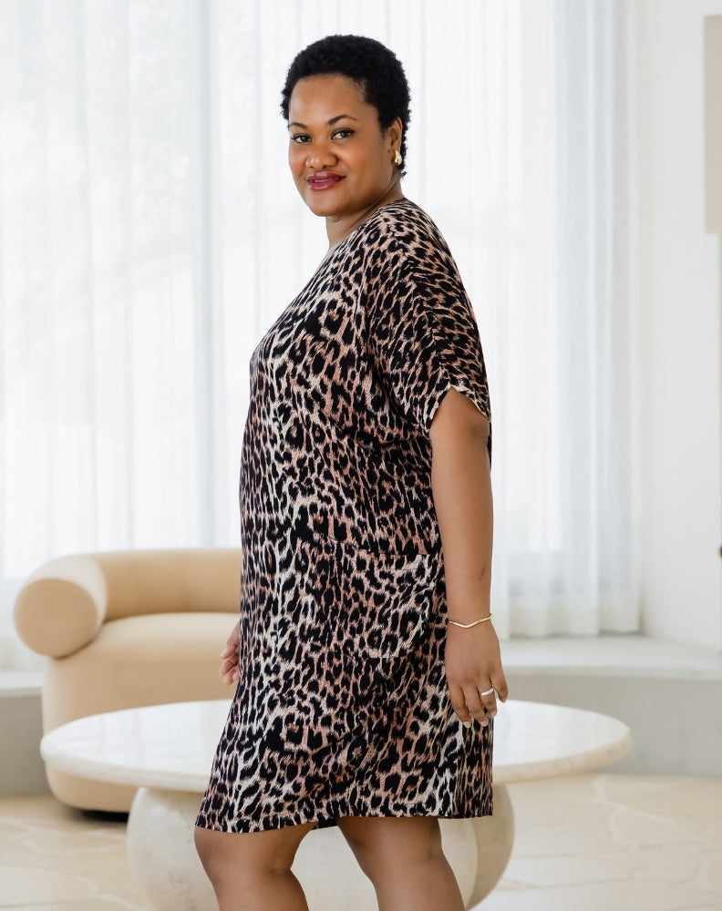 Woman wearing a leopard print dress in a room with white curtains and a beige chair.
