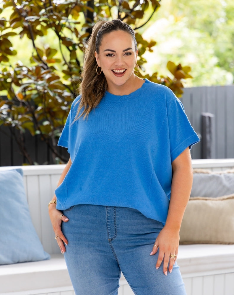 A woman wearing a bright blue short-sleeve knit top and blue jeans, standing outdoors in front of a white bench with pillows and green foliage.