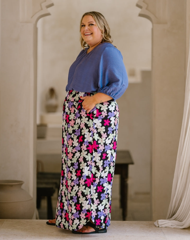 A woman modeling a two-piece outfit consisting of a dusty blue long-sleeve blouse and a high-waisted maxi skirt with a colorful floral pattern in pink, purple, white, and green, set against a warm, neutral-toned indoor background.