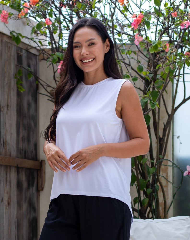 Woman wearing a white sleeveless top and black pants standing outdoors with plants in the background