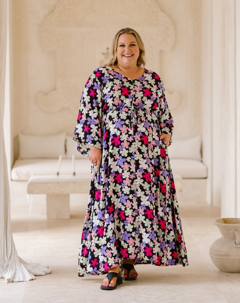 A woman wearing a long-sleeved, flowy maxi dress with a colorful floral pattern, standing in a bright, neutral-toned indoor setting.