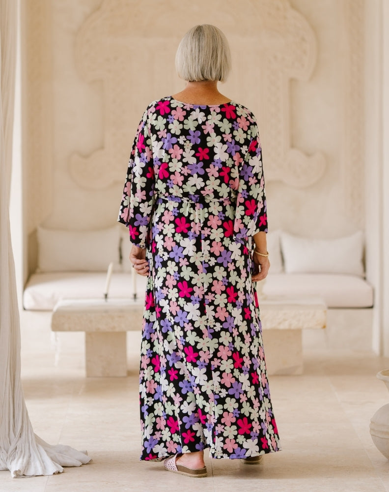 A woman viewed from behind wearing a long-sleeved maxi dress with a vibrant multicolor floral pattern on a black background, standing in a bright, neutral-toned indoor setting.