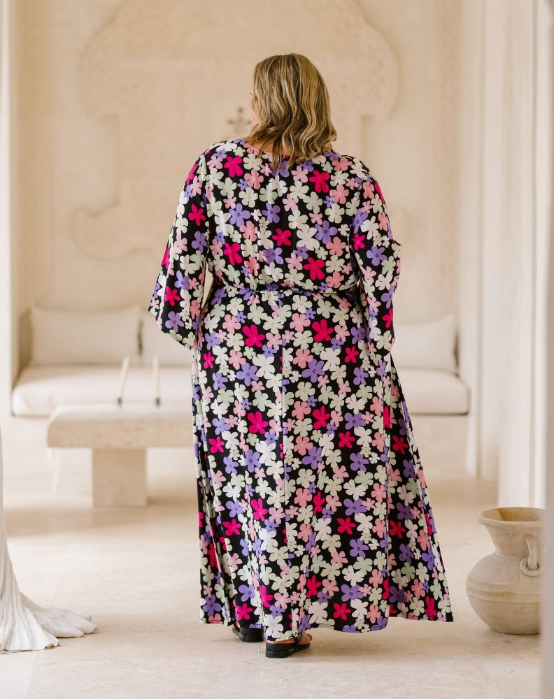 A woman viewed from behind wearing a long-sleeved maxi dress with a dense multicolor floral print on a dark background, standing in a bright, neutral-toned indoor setting.