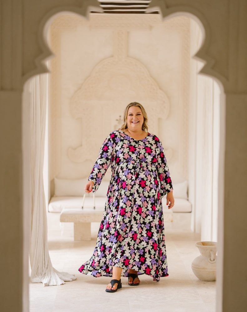 A woman wearing a long-sleeved maxi dress with a colorful floral pattern stands in a bright, cream-colored architectural setting framed by arches.