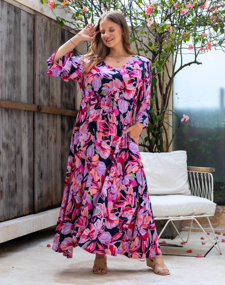 Woman wearing a colorful floral dress standing outdoors with a wooden wall and white chair in the background.