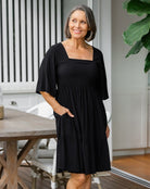 Woman wearing a black dress standing in a room with a wooden table and white cabinets.
