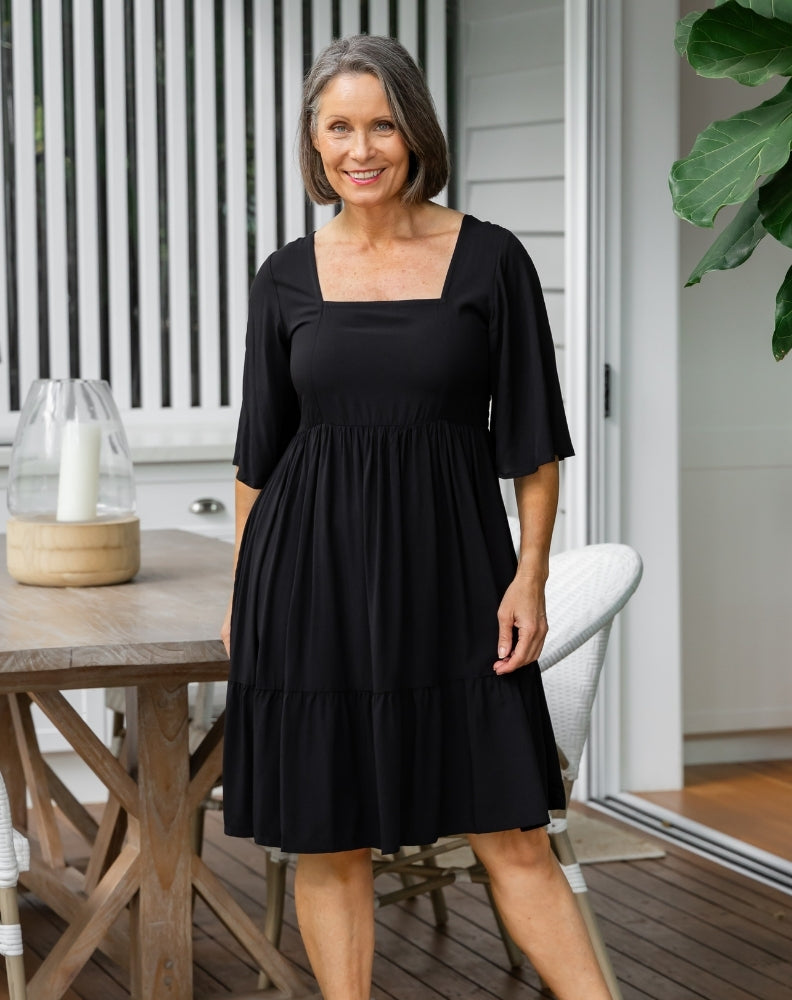 Woman wearing a black dress standing in a modern outdoor setting with a table and chairs.