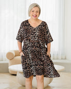 Woman wearing a leopard print dress in a room with white curtains and a beige chair.
